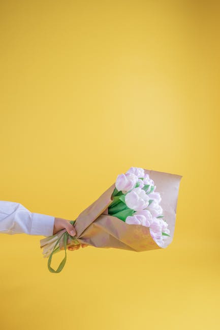 A person’s hand, wearing a white long-sleeve shirt, extending from the left side of the image, holds a bouquet of fresh, pale pink tulips with green stems and leaves. The bouquet is wrapped in light