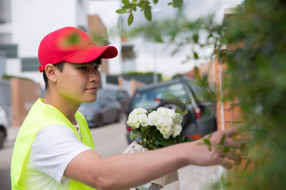 A young delivery person wearing a red cap and high-visibility yellow vest is carefully placing a floral gift of white hydrangeas into a designated spot on an outdoor wall. The arrangement features a l