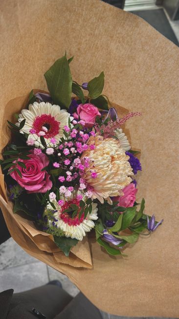 A vibrant bouquet of fresh flowers featuring pink roses, white daisies with pink centers, cream-colored carnations, and small purple flowers, arranged with green foliage. The flowers are wrapped in na