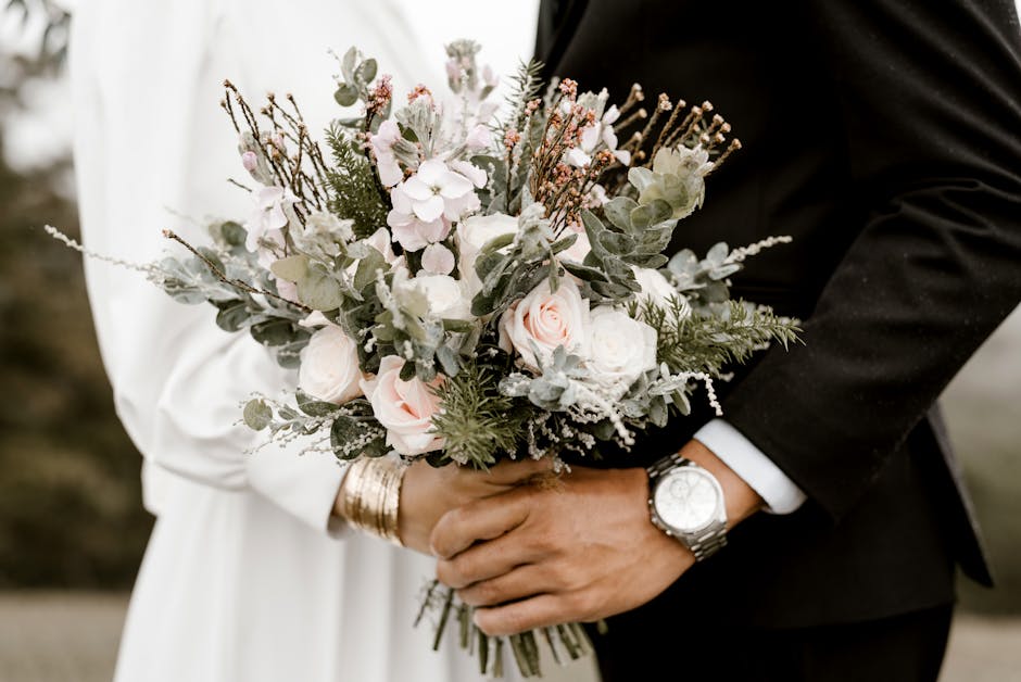 A person dressed in a white outfit holds a large bouquet of fresh flowers, including pale pink roses, white blossoms, and grey-green foliage such as eucalyptus, arranged in a loose, natural style. The