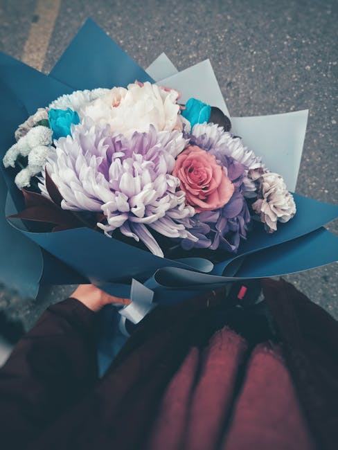 A hand holds a bouquet of fresh flowers wrapped in blue and white paper, featuring a large white chrysanthemum, a pink rose, purple lisianthus, and small blue accents. The flowers display vibrant colo