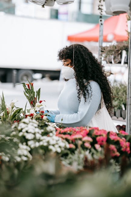 A woman with long, dark, curly hair wearing a white face mask and a light blue sweater is selecting fresh flowers at a florist's market stall. She holds a bouquet of red and white flowers, possibly li