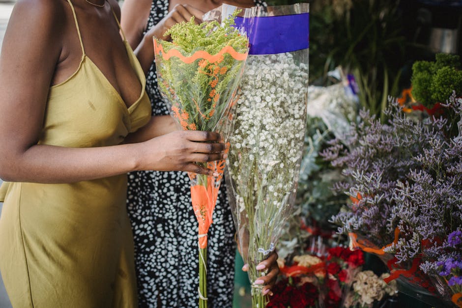 A woman in a yellow satin dress holds a tall bouquet of mixed green and pastel-colored flowers, including light green and lavender blooms, wrapped in clear floral cellophane with orange accents. She i